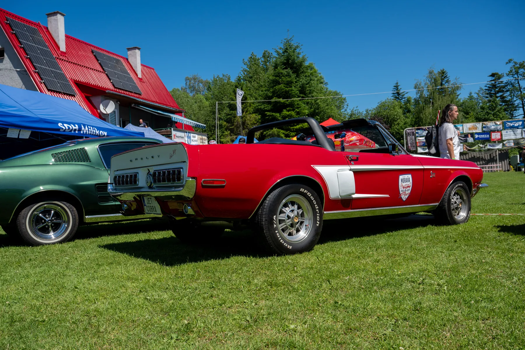 Červený Shelby GT500 kabriolet a zelený Ford Mustang fastback na trávníku areálu TJ Milíkov